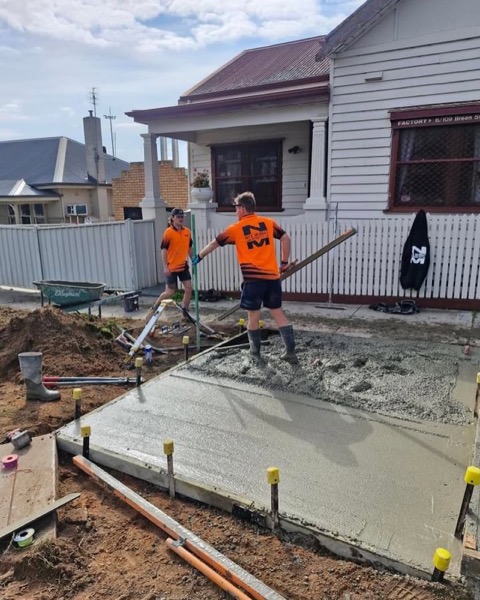 Two concreters in hi-vis screeding a fresh pour along the side of a weatherboard house