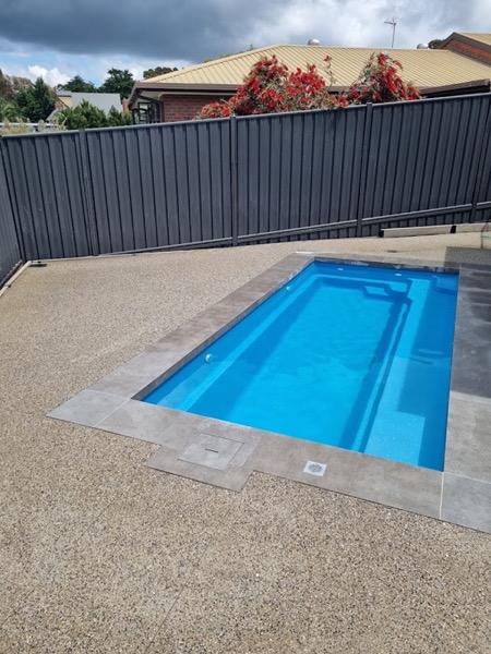 An exposed aggregate pool surround wrapping a blue fibreglass pool inside a colorbond fence