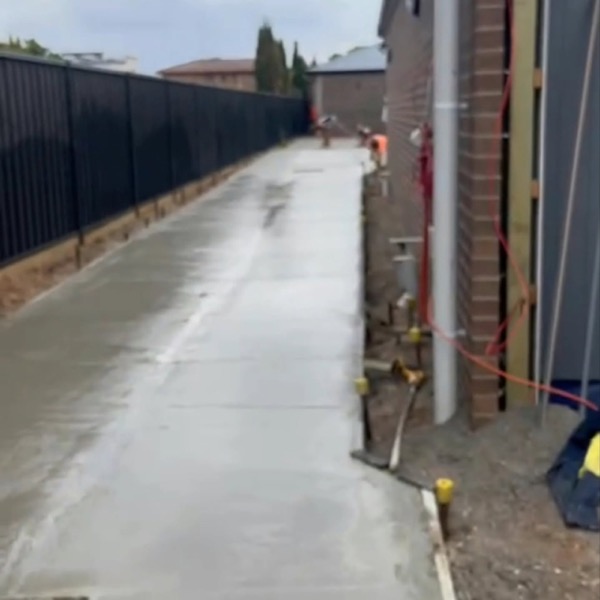 A concrete path running down the side of a brick house between the wall and a fence