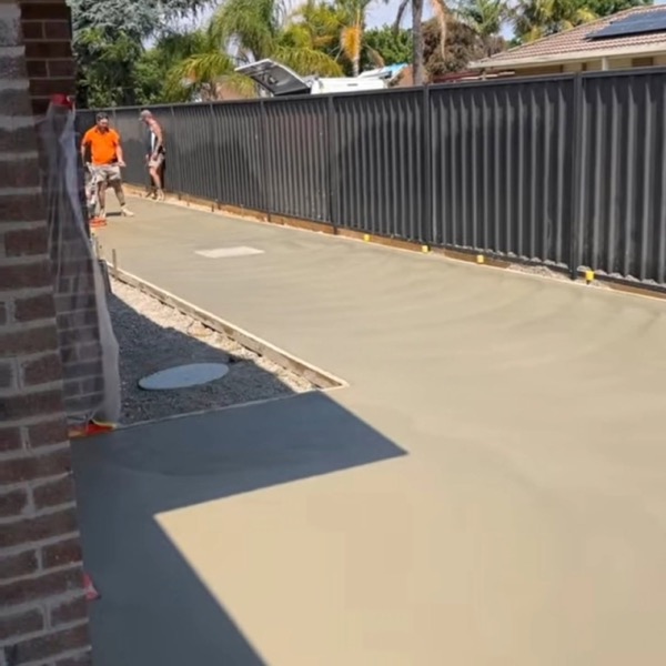 A freshly poured driveway strip between a brick wall and a colorbond fence, crew working at the far end