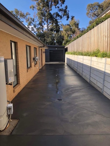 A charcoal coloured concrete driveway running between a brick house and a shed