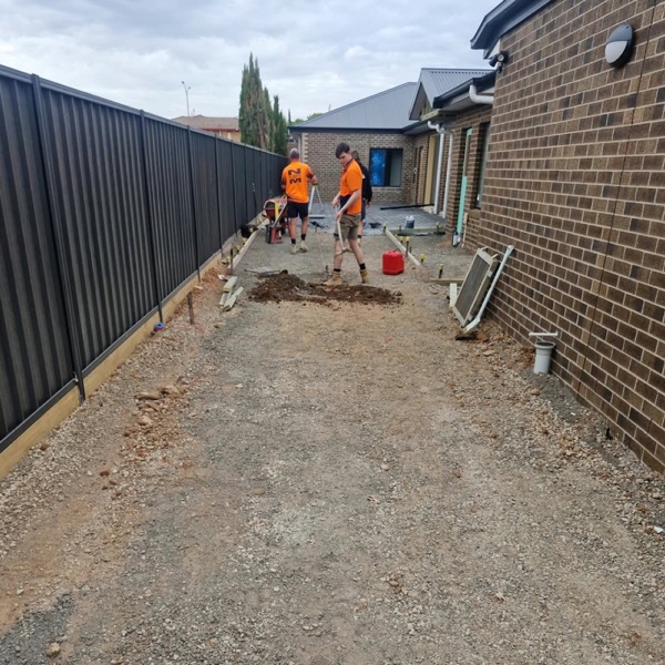 Two concreters in hi-vis preparing a narrow site between a brick house and a fence