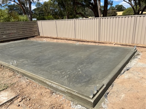 A finished shed slab with anchor bolts set, beside a colorbond fence and gum trees