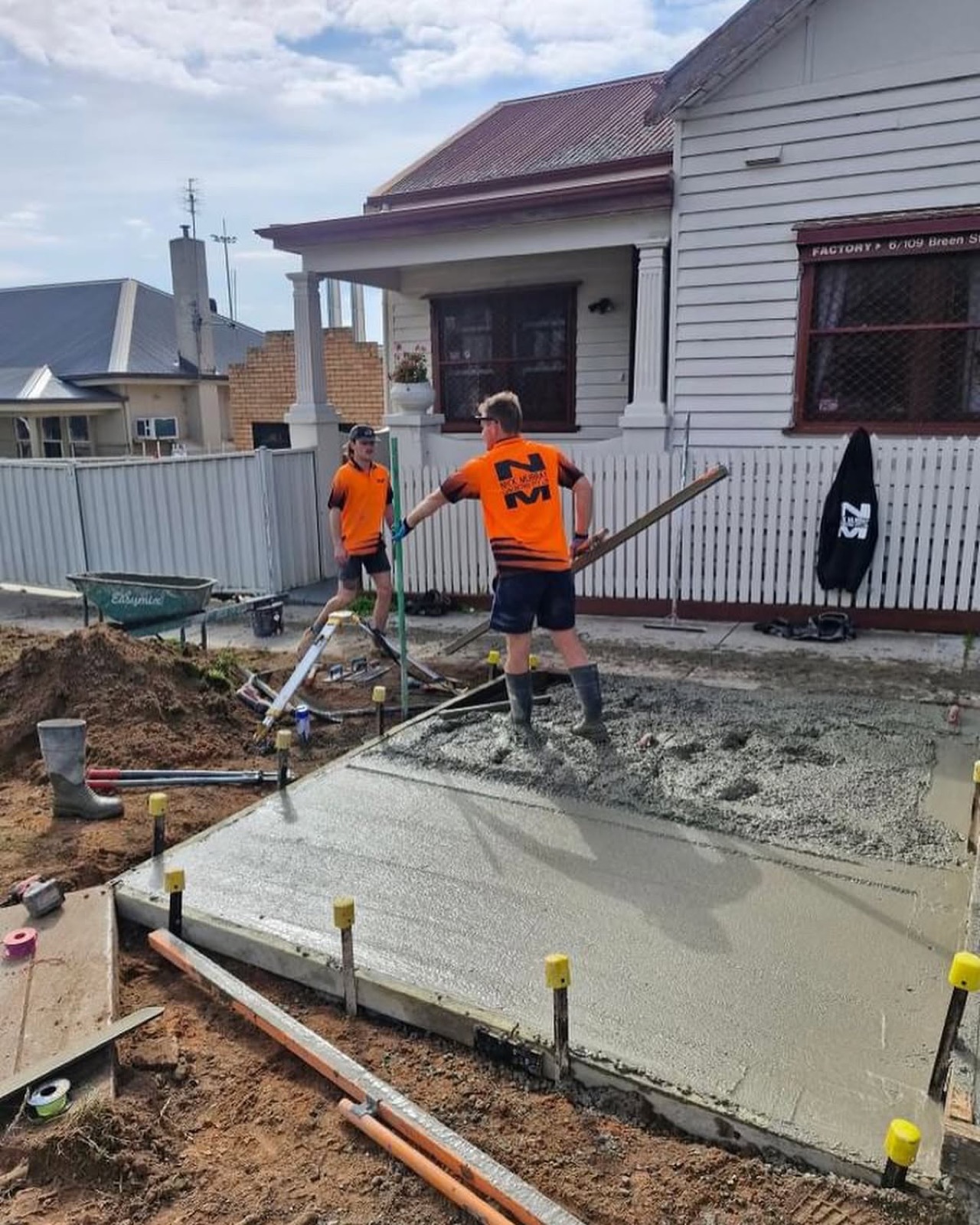 Two concreters in hi-vis screeding a fresh pour along the side of a weatherboard house