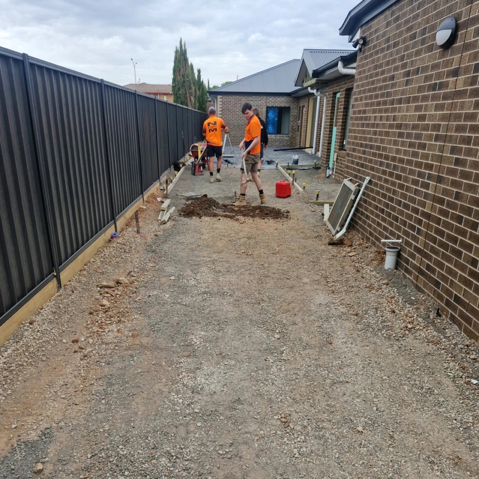 Two concreters in hi-vis preparing a narrow site between a brick house and a fence